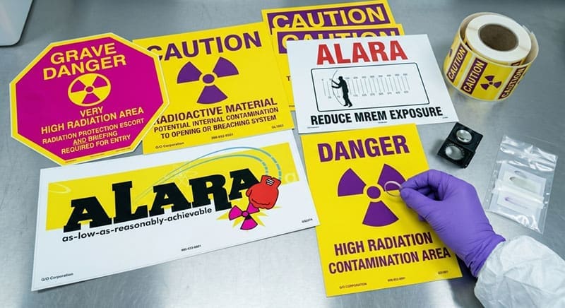 Composite quality control layout of specialized radiation safety labels, including 'Grave Danger,' 'Radioactive Material Caution,' and 'ALARA Reduce MREM Exposure' signs, being inspected by a gloved hand.